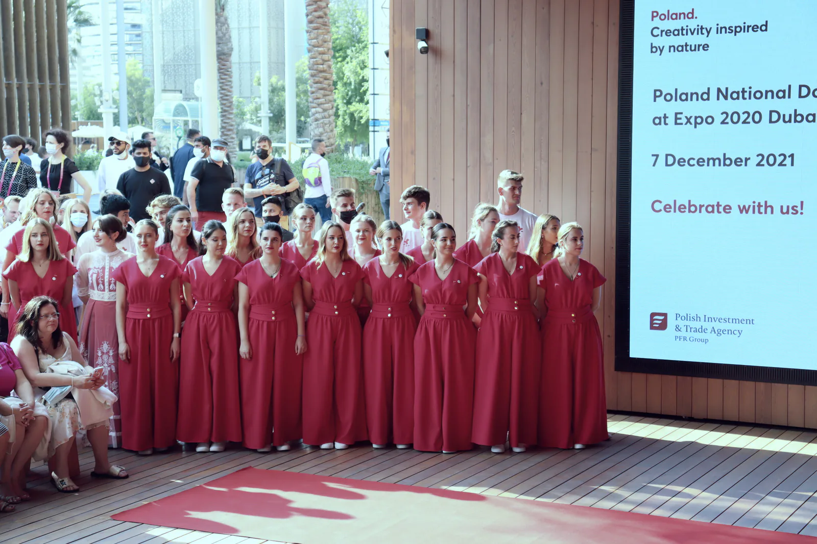 Young performers in red dresses lined up beside a Poland National Day at Expo 2020 Dubai screen dated 7 December 2021