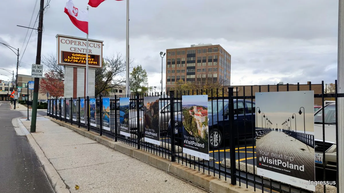 Your next trip #VisitPoland posters on the fence along the sidewalk by the Copernicus Center sign in Chicago, with a Polish flag above