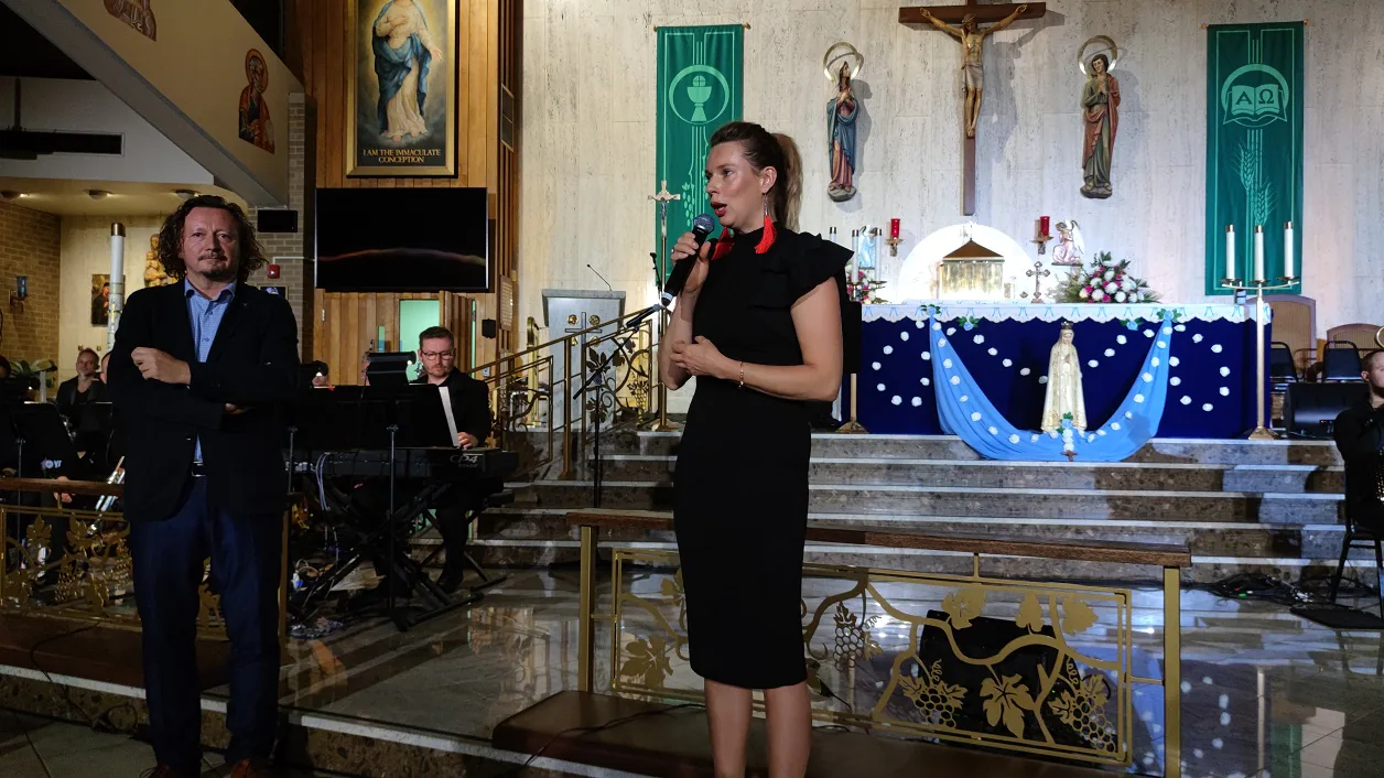 Performer in a black dress speaking into a microphone in front of the altar during The Saint concert, with musicians behind