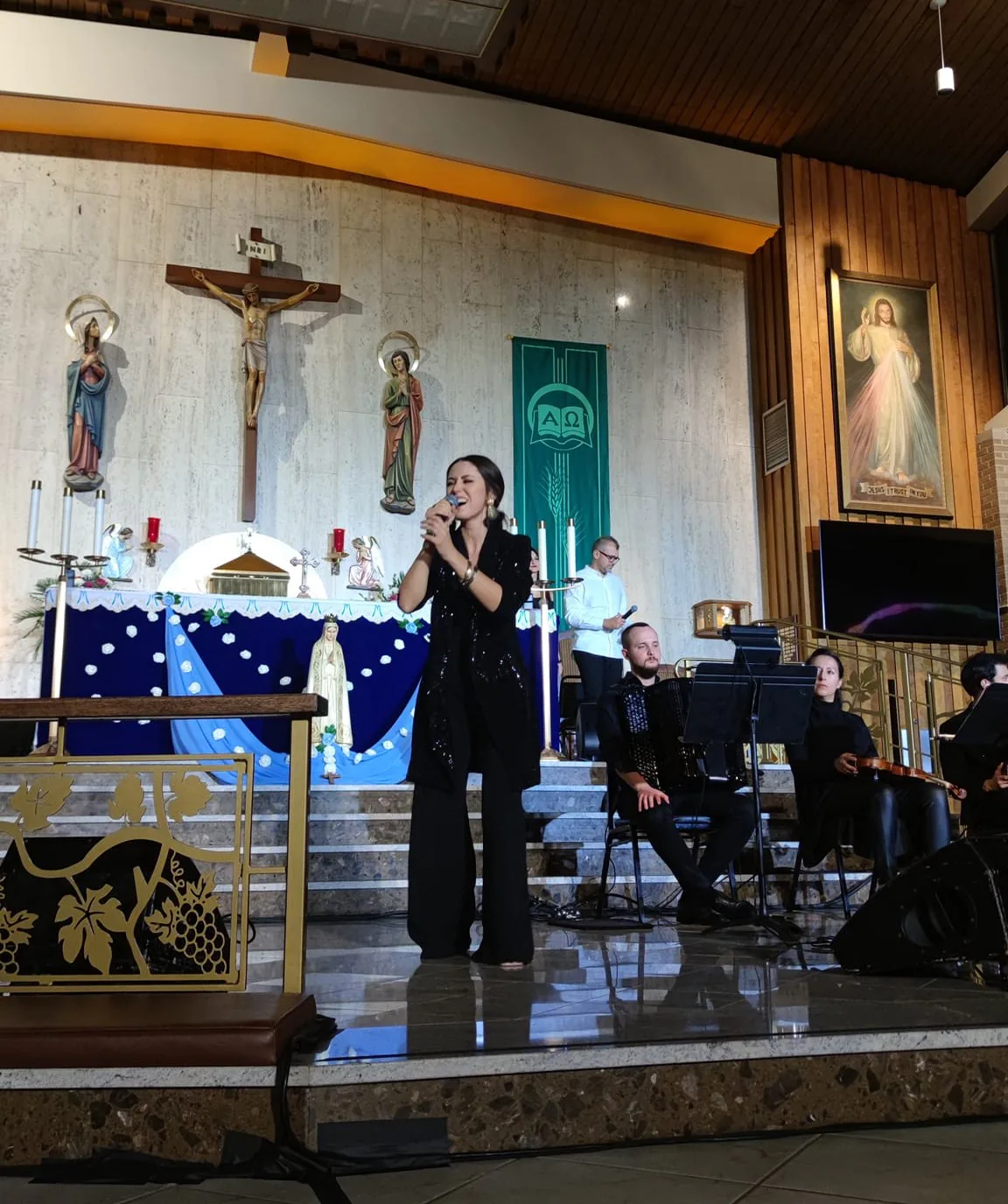 Singer in a sequined black outfit performing in front of the altar, with an accordionist and violinist behind her