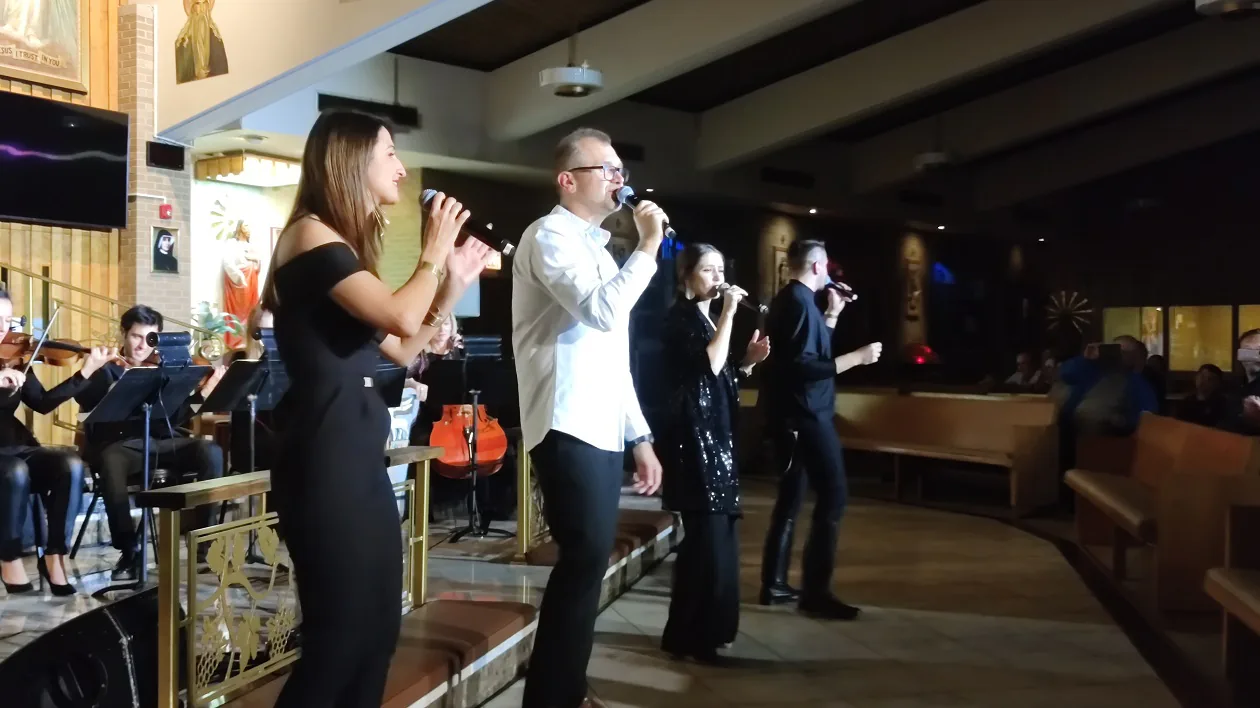 Four singers performing together with microphones beside the string section during The Saint concert in a parish church