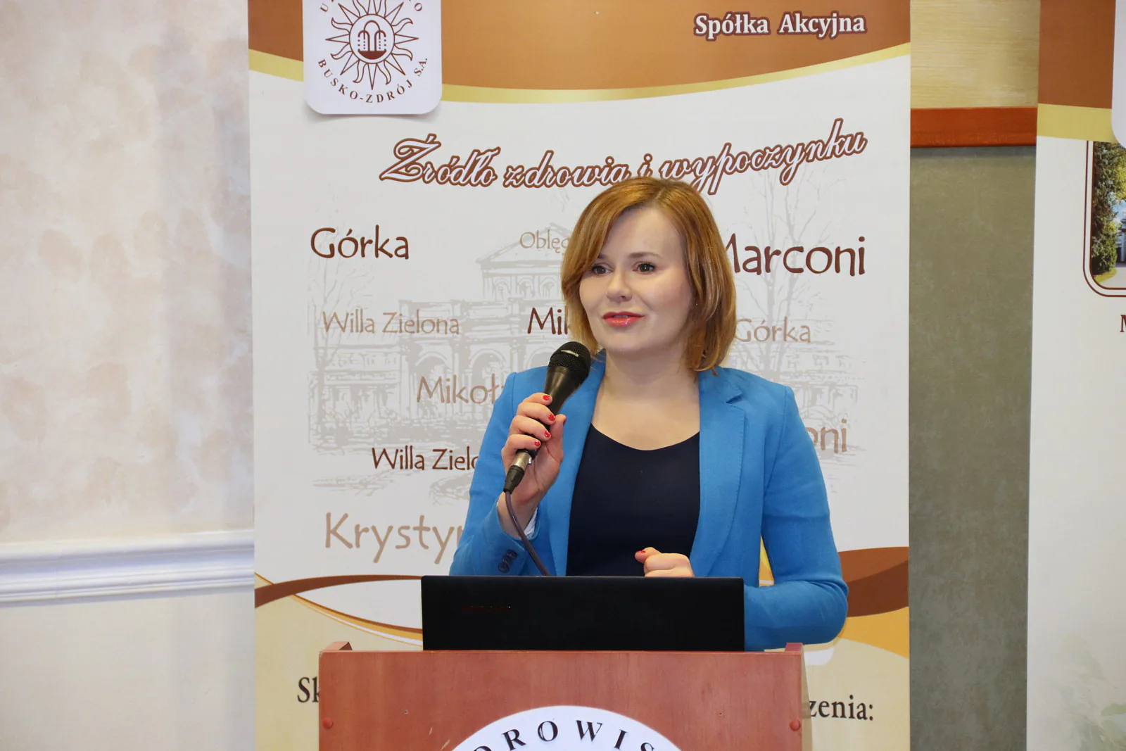 Speaker with a microphone presenting in front of Uzdrowisko Busko-Zdrój banners during the 2019 study tour of Polish spas
