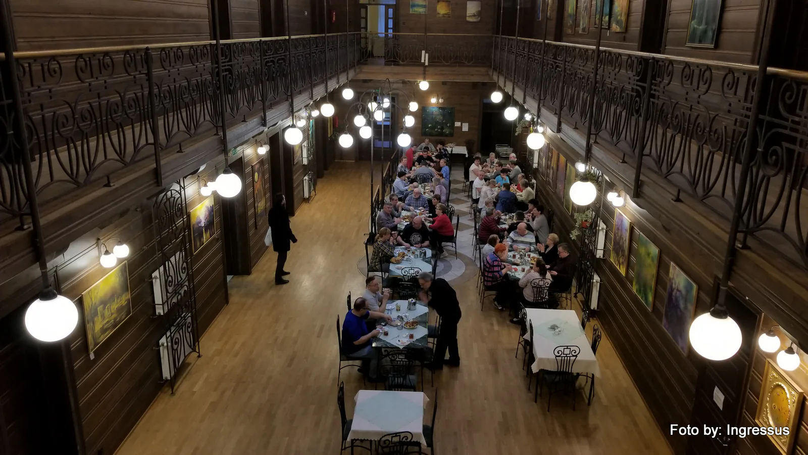 Guests at long tables in a wood-paneled dining hall with wrought-iron balconies and globe lamps at a Polish spa