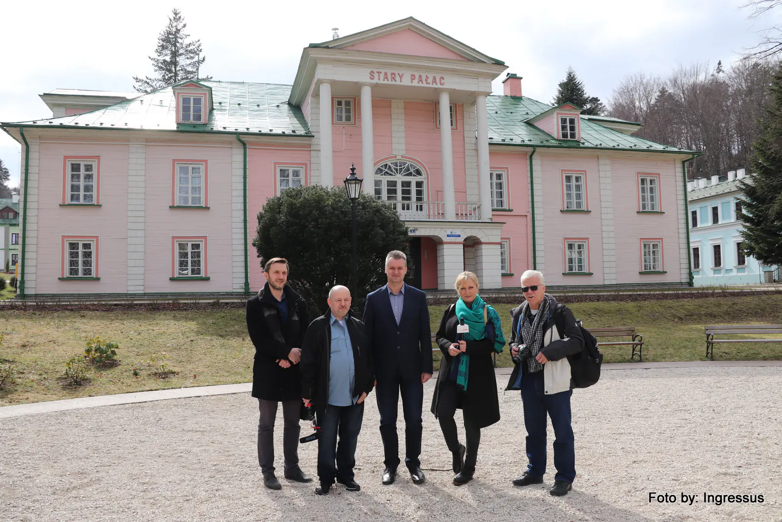 Study tour group with a reporter holding a microphone in front of the pink Stary Pałac building in a Polish spa town