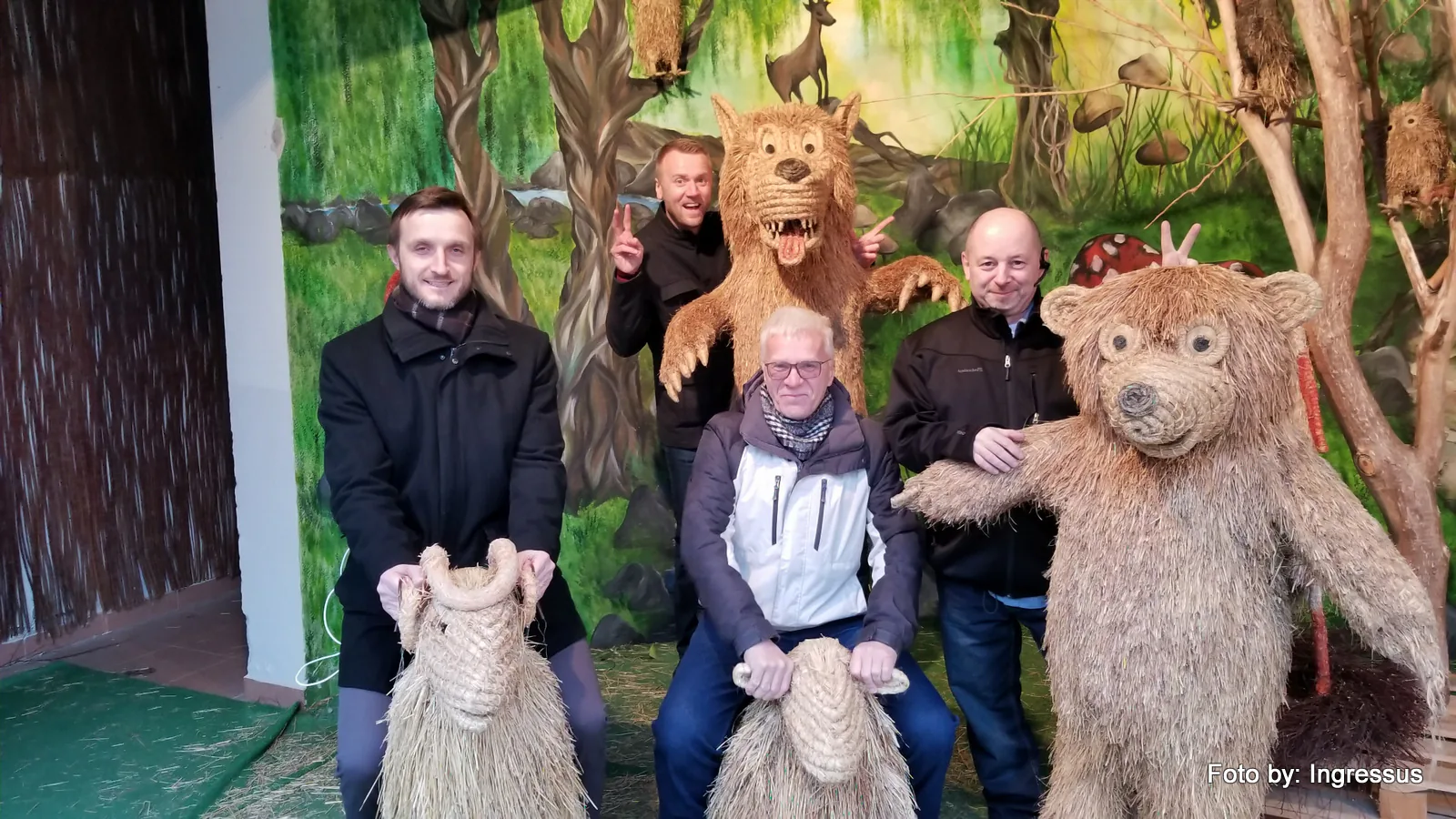 Four men posing with large straw figures of a bear, a lion and goats in front of a forest mural, 2019 study tour