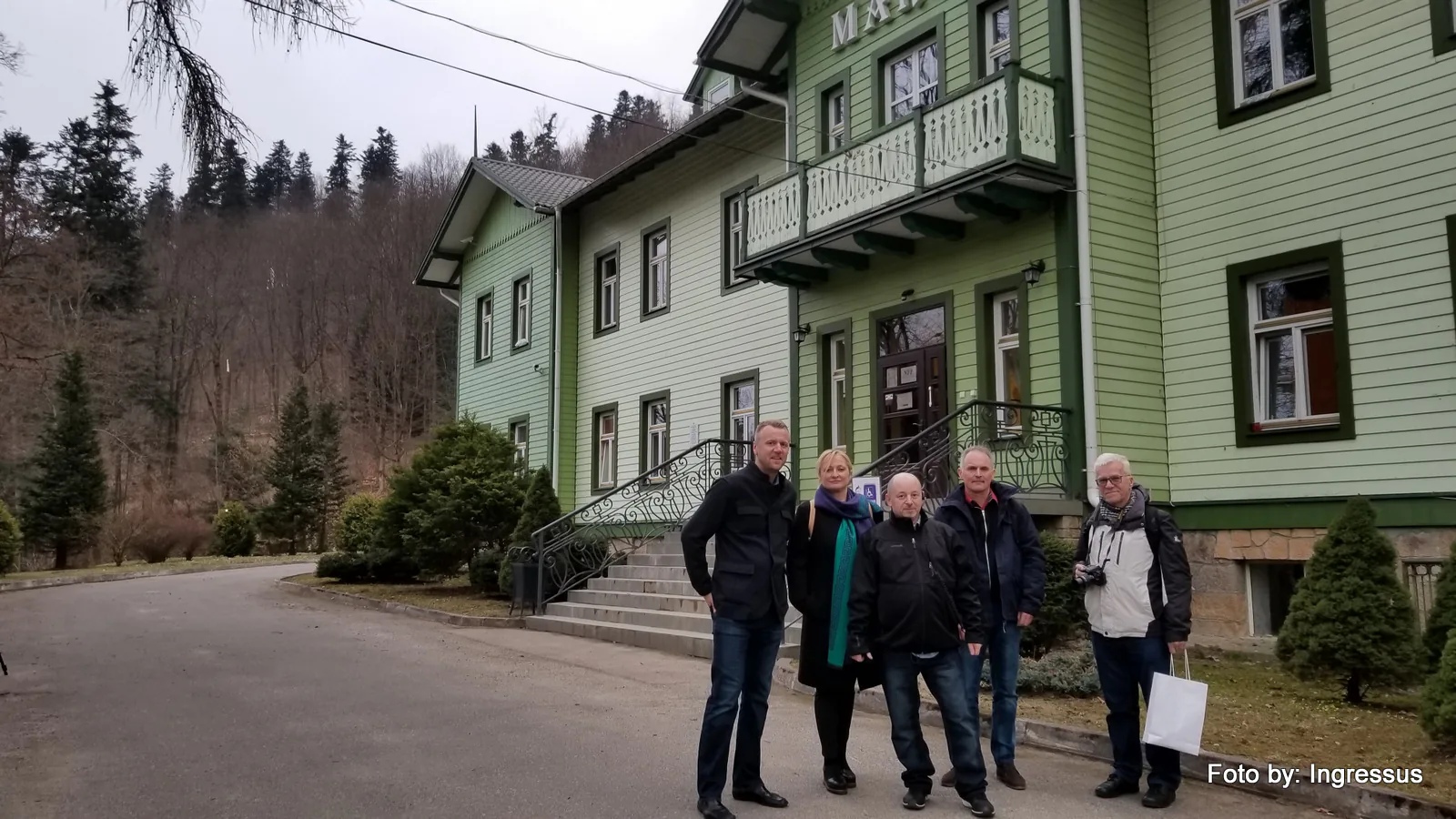 Study tour group in front of a green wooden villa with a carved balcony below forested hills in a Polish spa town
