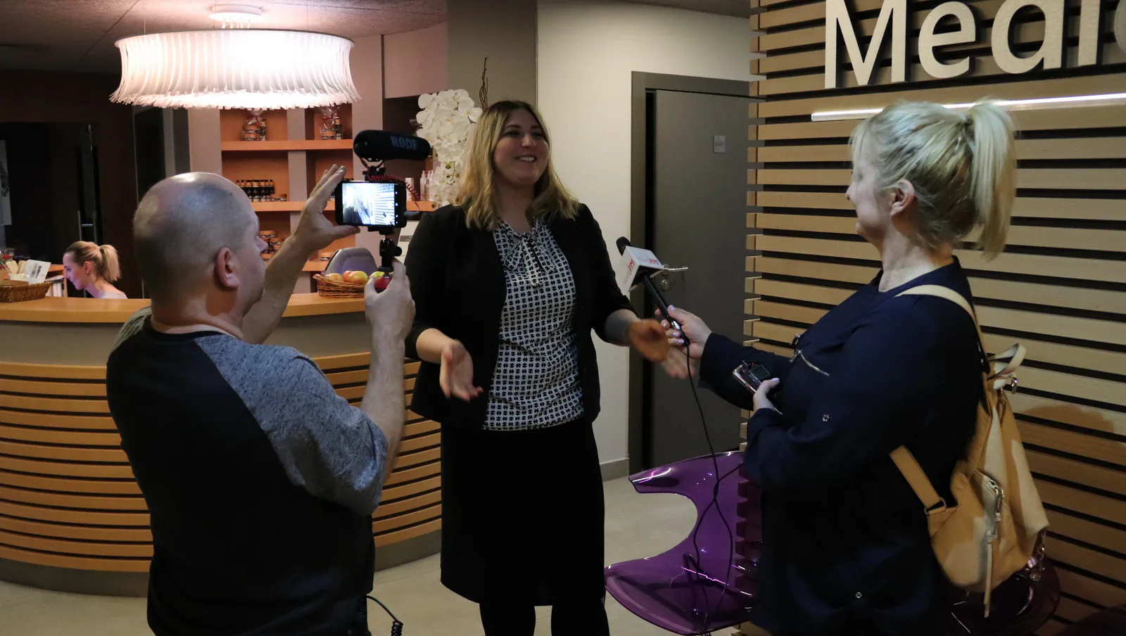 Journalist interviewing a woman with a microphone while another participant films on a phone in a spa reception area, Poland