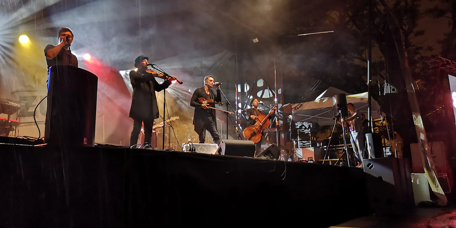 Zakopower performing at night on the Taste of Polonia stage in the rain, with violins and a cello under colored stage lights